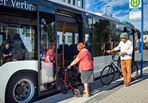 &Auml;ltere Frau mit Rollator steigt in einen Linienbus an einer Haltestelle ein, Mann mit Fahrrad wartet daneben.